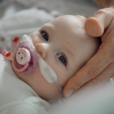 Toddler with nasal oxygen prongs insitu lying with head on pillow and sucking on dummy. Adult hand supporting head.